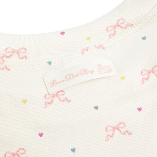 Close-up detail of the Bow Print Cropped T-Shirt showcasing the Lana Del Rey label and ivory fabric printed with delicate pink ribbon bows and pastel heart motifs, highlighting the soft vintage-inspired pattern.