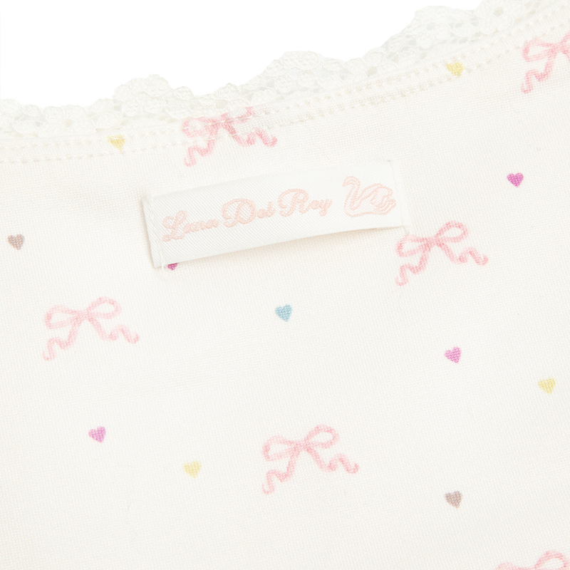 Close-up detail of the Bow Print Cropped Cami Tank Top showcasing the delicate lace trim, Lana Del Rey label, and ivory fabric printed with soft pink ribbon bows and pastel heart motifs.
