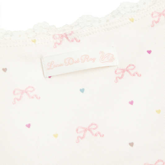 Close-up detail of the Bow Print Cropped Cami Tank Top showcasing the delicate lace trim, Lana Del Rey label, and ivory fabric printed with soft pink ribbon bows and pastel heart motifs.
