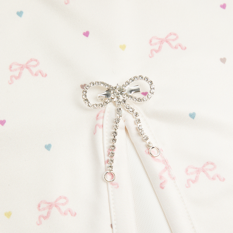 Close-up detail of the Bow Print Mini Skirt showcasing the sparkling rhinestone bow charm with adjustable drawstring on ivory fabric printed with delicate pink ribbon bows and pastel heart motifs.