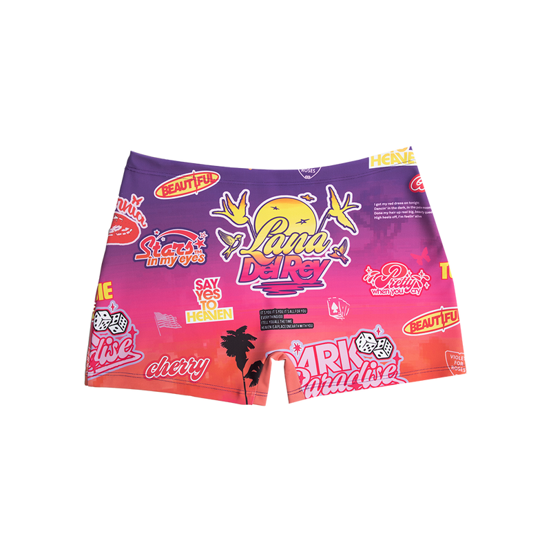 Front view of the Paradise Print Biker Shorts featuring an all-over sunset gradient with Lana Del Rey logo, retro stickers, palm trees, cherries, dice, birds, and Paradise era inspired graphics in vibrant pink, purple, and orange tones.
