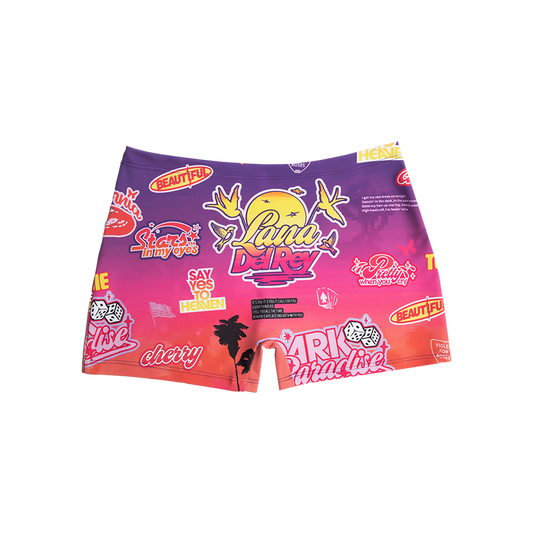 Front view of the Paradise Print Biker Shorts featuring an all-over sunset gradient with Lana Del Rey logo, retro stickers, palm trees, cherries, dice, birds, and Paradise era inspired graphics in vibrant pink, purple, and orange tones.